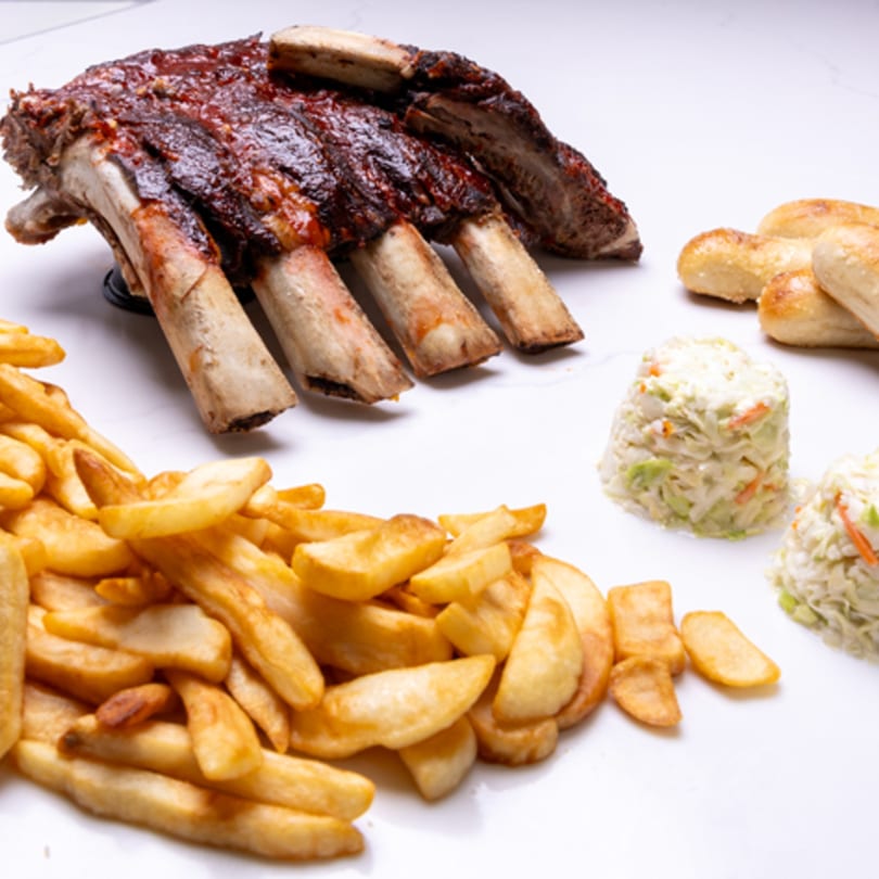 Barbecue ribs with french fries, coleslaw, and breadsticks on a white surface.